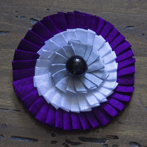 a cockade with three different narrowly pleated ribbons in purple, white and grey, plus a shiny black bead in the middle.