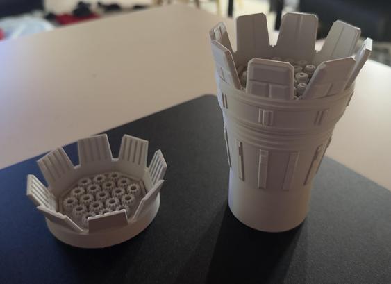 The image features two white 3D-printed objects on a dark surface. The larger object resembles a tall, cylindrical container with a detailed top, while the smaller object is a shorter, wider bowl-like shape with decorative elements around the edge. Both