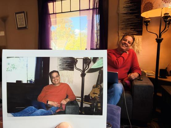 A devastatingly handsome man sitting on a couch, and a hand holding up a photo of that same guy on a couch.
