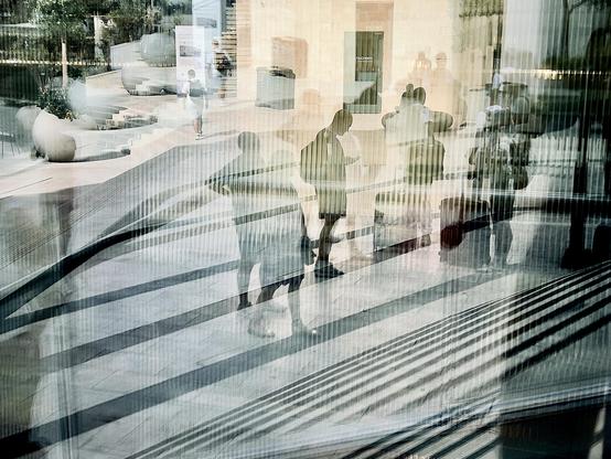 In a busy modern square people come and go and are seen in the reflection of a plate-glass panel walking through patches of bright sunlight and shadow. The exposure chosen turns them into silhouettes.