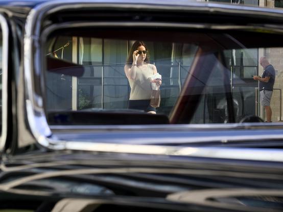 A young woman wearing sunglasses and carrying her cellphone and food in a paper bag is framed through the rear and front windshields of a classic pickup truck parked in an open square. In the background a man glances at his cellphone.