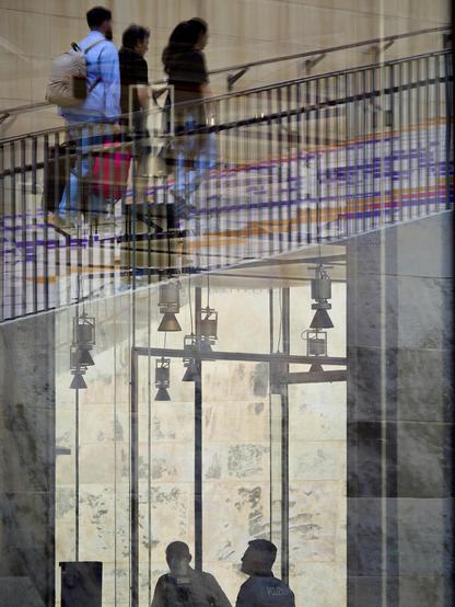 At first glance it is hard to tell what is happening in this scene as three people dragging luggage up a flight of stone steps appear high above the silhouettes of two men. The people on the steps are a reflection of what is happening behind the camera, made on the tall glass building in which the two men are working.