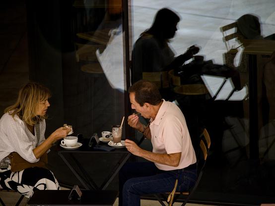 A couple drinking fancy coffees and eating desserts sit at an outside table at a cafe while their shadows are cast onto a dark plate-glass window, creating a perfect shadow reproduction of the scene.