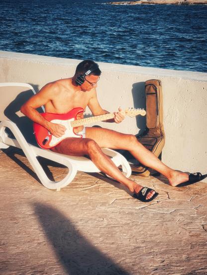 A man sat on a lounger by the pool and sea plays a red electric guitar while wearing headphones, swimming trunks and pool sliders. I wondered if he was practising House of the Rising Sunbed?