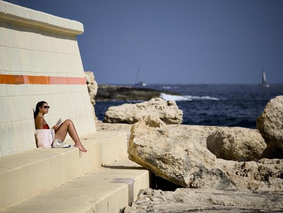 A young woman sits alone at the end of a concrete breakwater, with her back against the wall and her knees raised. She is wearing sunglasses, a swimsuit and has a book open in her lap. A bag and her shoes are by her side. She is staring out to sea across the rocky coastline.