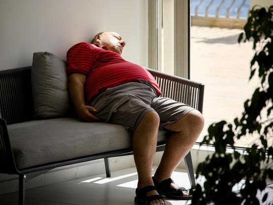 A man wearing a red short-sleeved shirt, shorts and sandals slumps in a chair in a hotel lobby. He is fast asleep in the warmth of the sunlight pouring through the full-length window next to him.