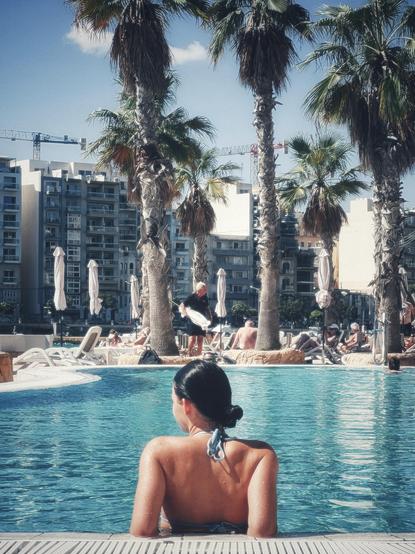 A young woman rests in the shallow end of a swimming pool with her elbows resting on the edge of the pool as she soaks up the warm sunshine. We see her from behind and she is looking off slightly to her left. Her dark hair is in a bun and her bikini is the same colour as the water in the pool, which is lined with tall palm trees. In the distance there are modern buildings across the bay and cranes at work.