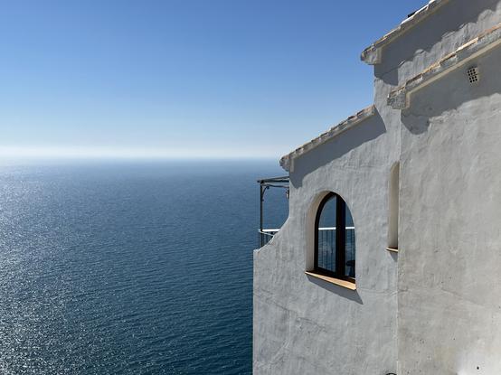 Eine weiss getünchte Wand mit einem halbrunden Fenster ragt hoch über einer Klippe übers Meer. Das blaue Wasser tief unten wird seitlich von der Sonne beschienen und verschwimmt am Horizont in einem hellen Dunststreifen mit dem Himmelsblau.