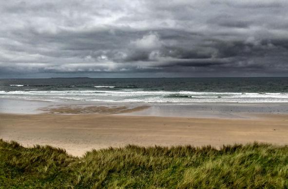The view stretches from the dunes in the foreground, covered in green dune grass, across the wide, empty, pale sandy beach and the shallow, white-capped surf, out over the dark sea to the horizon, above which dense, dark-grey storm clouds have gathered. 

Der Blick geht über die mit grünem Dünengras bewachsenen Dünen im Vordergrund über den leeren breiten, hellen Sandstrand und die flache weißschaumige Brandung über das dunkle Meer bis zum Horizont, über dem sich dichte dunkelgraue Gewitterwolken gebildet haben.
