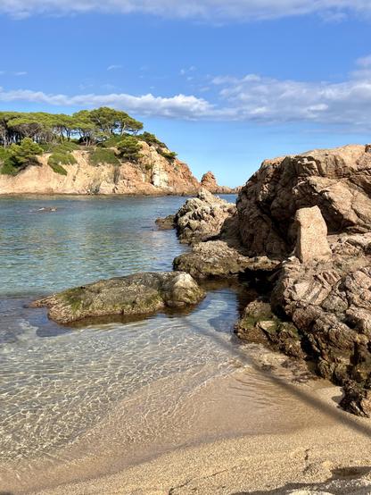 A serene coastal scene featuring rocky formations and clear water. Green trees are visible on a cliff in the background, under a partly cloudy blue sky. The sandy beach is in the foreground, with gentle waves lapping against the rocks.

Eine ruhige Küstenszene mit Felsformationen und klarem Wasser. Grüne Bäume sind auf einer Klippe im Hintergrund zu sehen, unter einem teilweise bewölkten blauen Himmel. Der Sandstrand steht im Vordergrund, mit sanften Wellen, die gegen die Felsen wischen.