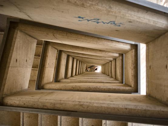 Looking down the centre of multiple flights of stone steps to people passing through a tiny space at the bottom in an image which has a disorienting feeling