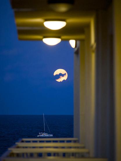 A small yacht sails beneath a bright moon just after sunset and is captured through the gap created by a series of balconies which are themselves lit by half-dome white lights.
