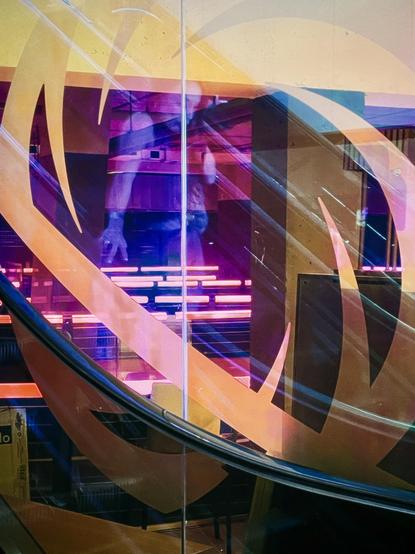 The reflection of a woman in the glass of an escalator can be seen as she descends into the neon-lit interior of a basement bowling alley