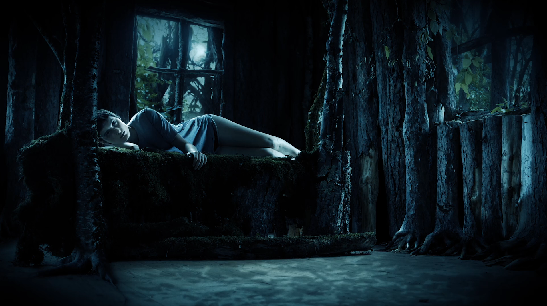 A screenshot from the film Mother of Flies. A young woman lies in her side in a bed of stone or wood and moss. It is nighttime and the scene is saturated a surreal blue. Moonlight spills in from a window behind her, framing her with a halo of light.