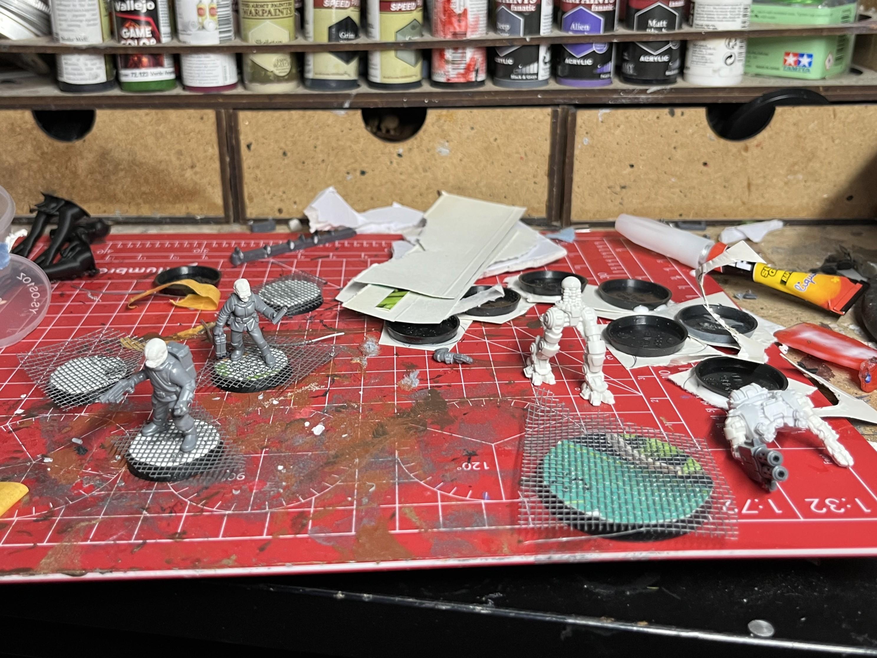 A messy hobby table with some unpainted 28mm models and some bases with a grating effect applied.