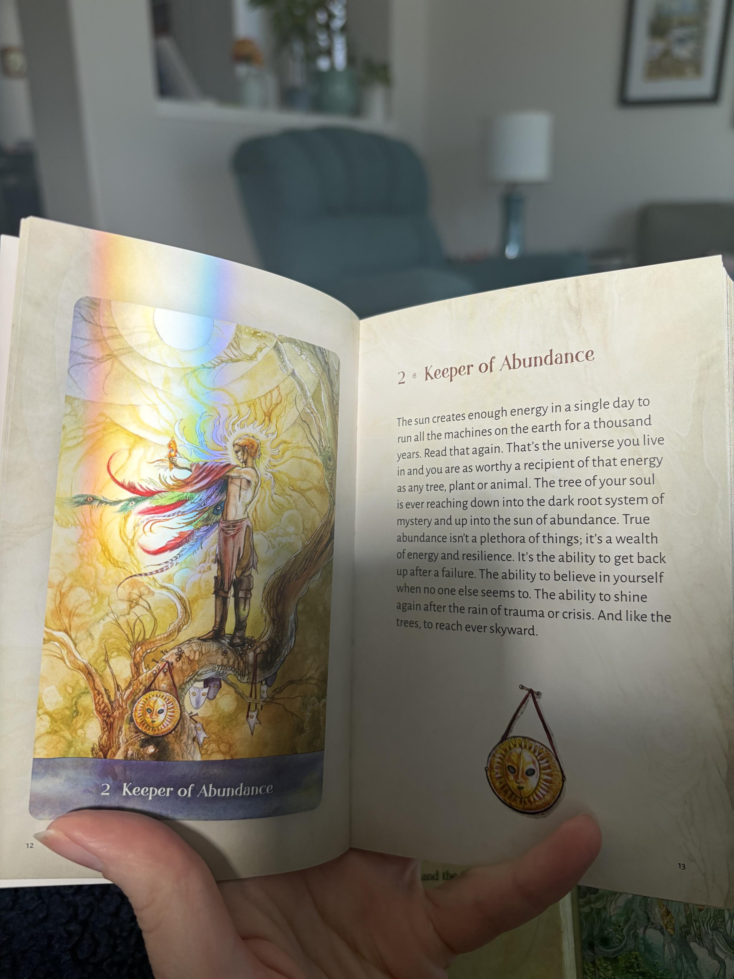 A page from the Tree Keepers Oracle book depicting a man standing on a tree branch bathed in sunlight, holding up a mask he’s taken off. There is a round sun mask at his feet that is repeated on the opposite page. An actual rainbow is shining on the artwork from a nearby window. The text reads: 

2 • Keeper of Abundance
The sun creates enough energy in a single day to run all the machines on the earth for a thousand years. Read that again. That's the universe you live in and you are as worthy a recipient of that energy as any tree, plant or animal. The tree of your soul is ever reaching down into the dark root system of mystery and up into the sun of abundance. True abundance isn't a plethora of things; it's a wealth of energy and resilience. It's the ability to get back up after a failure. The ability to believe in yourself when no one else seems to. The ability to shine again after the rain of trauma or crisis. And like the trees, to reach ever skyward.