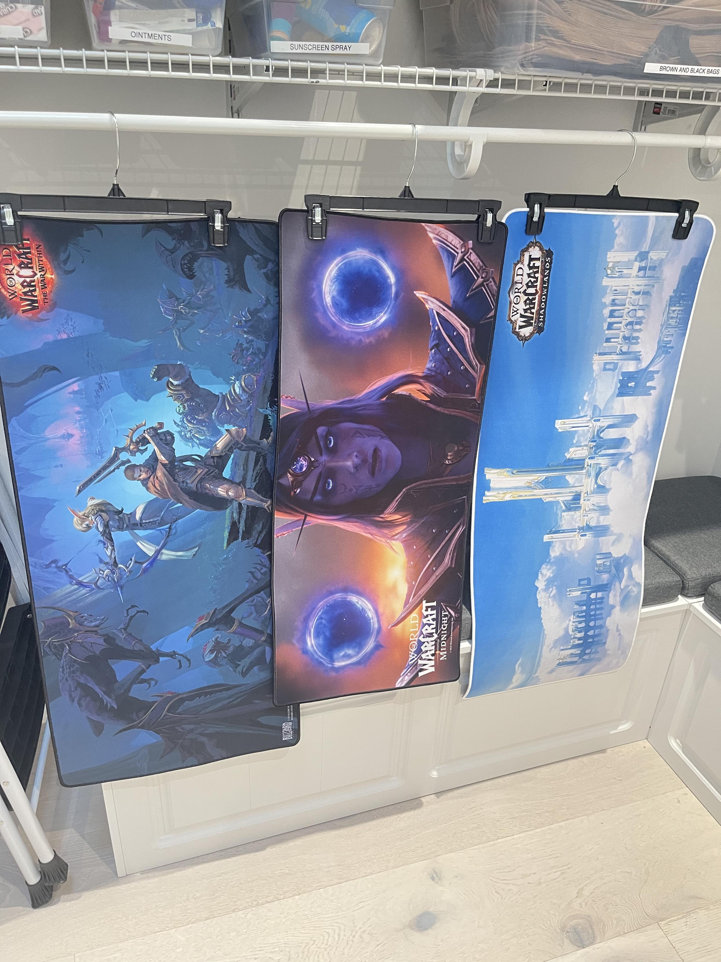 A photo of three gaming desk mats. On the far left is the World Within Key art, featuring Anduin, Thrall, and Alleria. In the middle hangs a mat featuring a head shot of Xal’atath with big purple void globes hovering above her shoulders. And finally on the right is a picture of of Bastion from Shadowlands, with its tall white towers floating in the clouds against a blue sky.