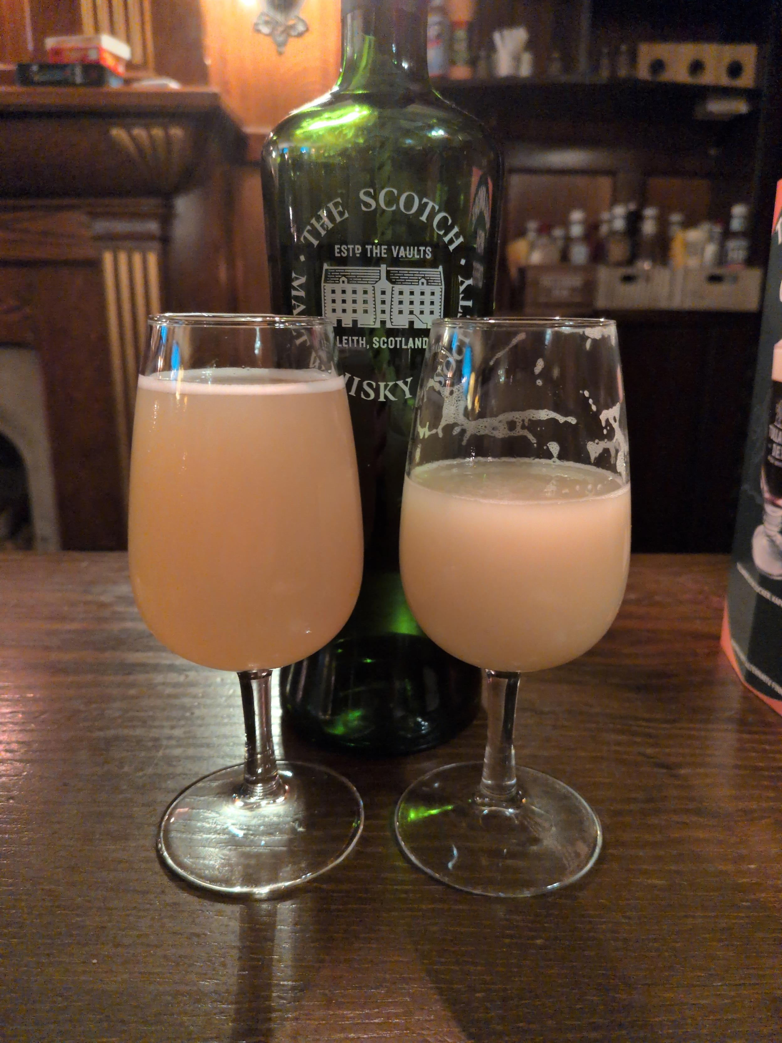 A photo of two small glasses of beer in front of a dark green whisky bottle. The beers are orange, the table they're on is dark brown. There is a shelf with stuff, and an open fireplace (no fire) in the background.
