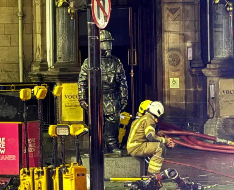 Photograph of two firefighters sitting on the plinth of the Citizen Firefighter statue outside the Grand Central Hotel. The statue is a tribute to fallen firefighters, designed by Kenny Hunter.