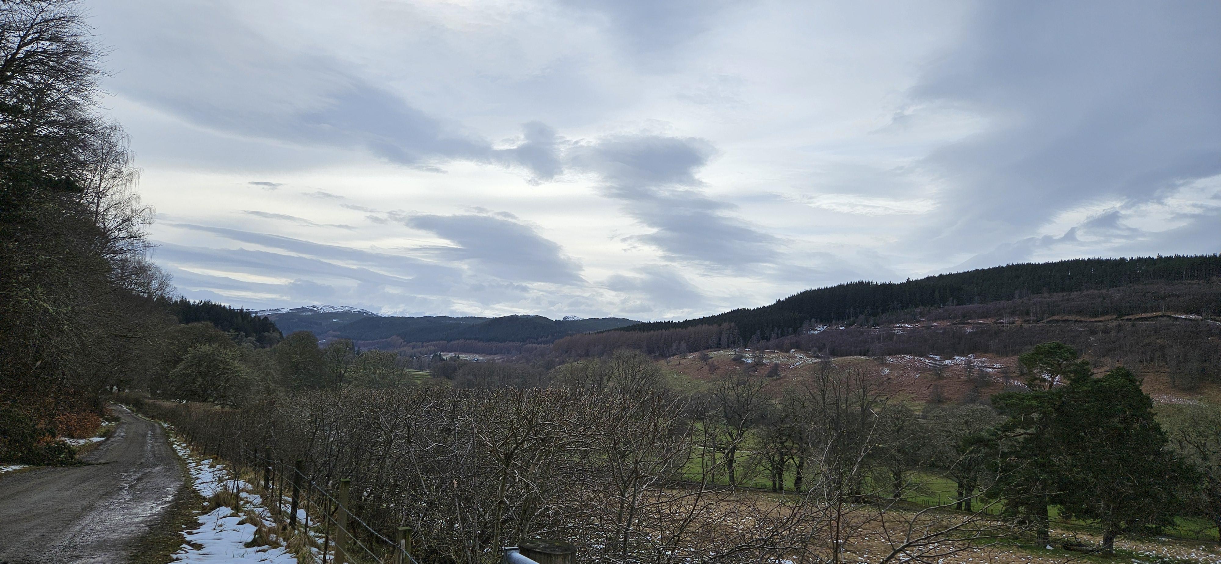 A gravel road winds along the left edge of a rural landscape, bordered by a wire fence and patches of snow. In the foreground, a field of leafless, thin-stemmed bushes is visible, with a wooden fence running along the bottom right. Rolling hills covered in a mix of bare trees and dark evergreens rise in the mid-ground, sloping down into a valley. In the far distance to the left, faint mountains with snow-capped peaks are visible against the horizon. The sky is overcast with a thick layer of grey and white clouds covering the upper portion of the image.

Provided by @altbot, generated privately and locally using Qwen3.5:9b

Manually Checked for good enough accuracy. 

🌱 Energy used: 1.791 Wh