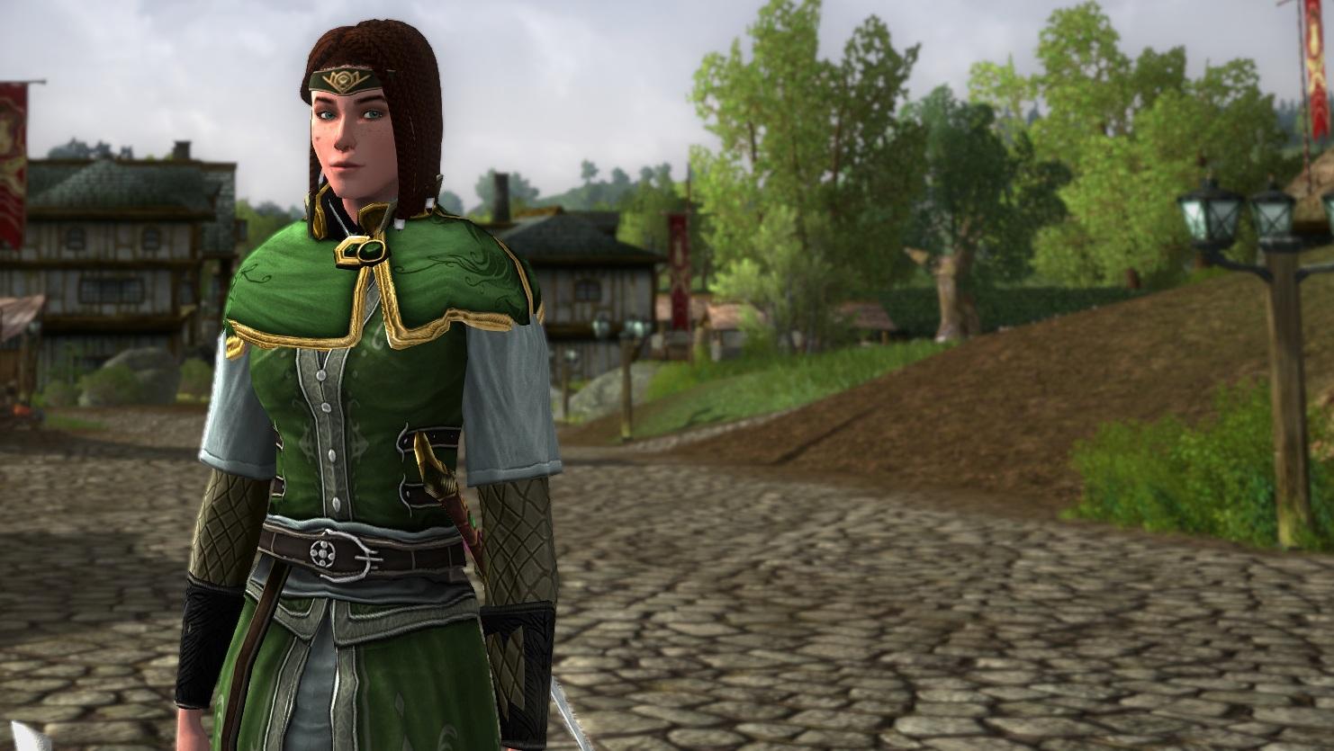 My main LOTRO character in a simple mid-green tunic with leather belt and a brighter green shoulder wrap. she's stood at a slight off-angle to the camera, and looking slightly sideways, directly at the camera. The picture is on the cobbled street of Bree, with trees as Middle-ages style wooden buildings blurred in the background.