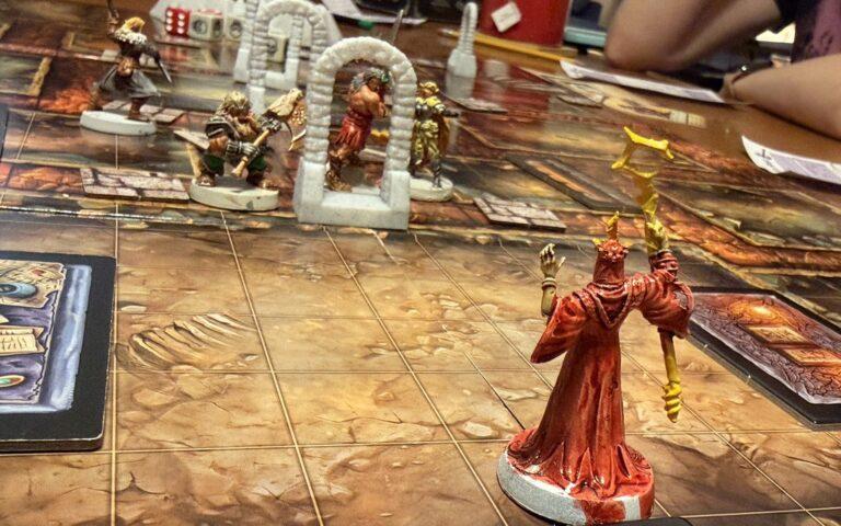 A HeroQuest First Light adventure in progress; a red-robed wizard mini is in the foreground; in the background, through a white arch, player minis are seen.