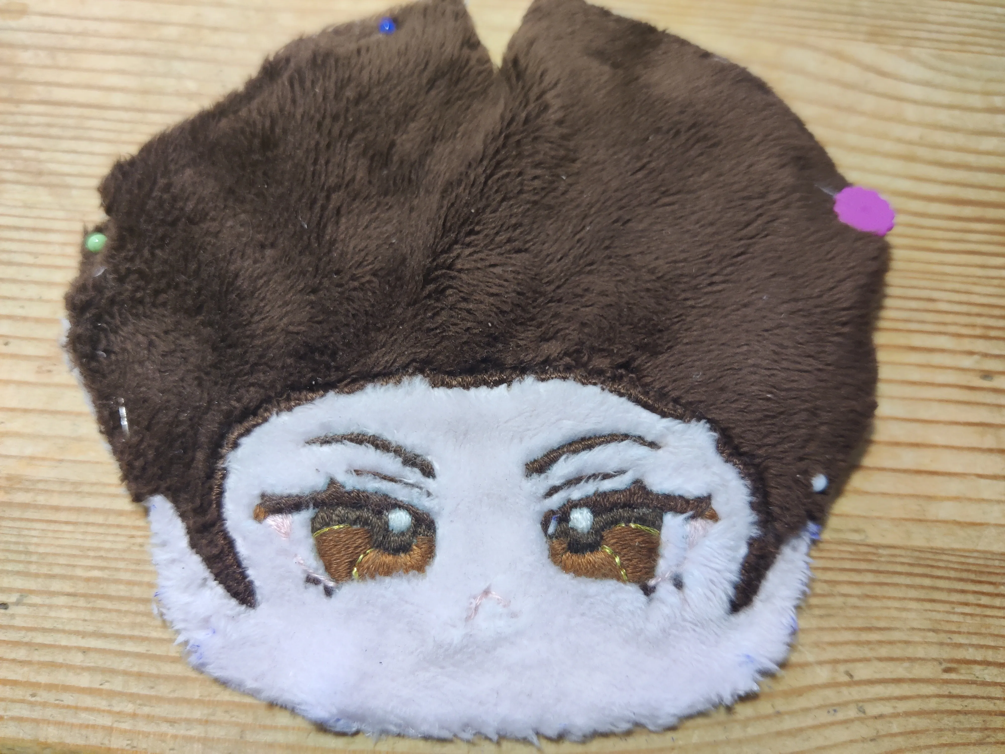 A fabric doll face laid out flat on a desk. A basic hairline has been embroidered, as well as "anime" style eyes and a frowny mouth.